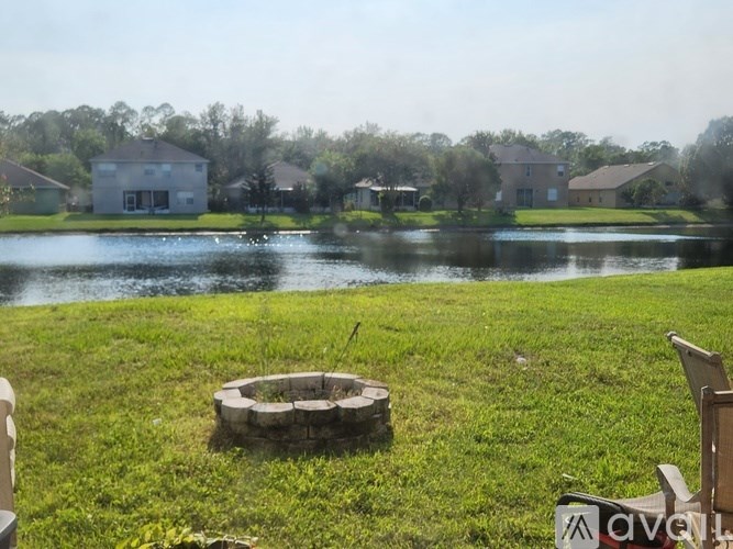 A fire pit sits in the middle of a grassy field with a lake and houses in the background.