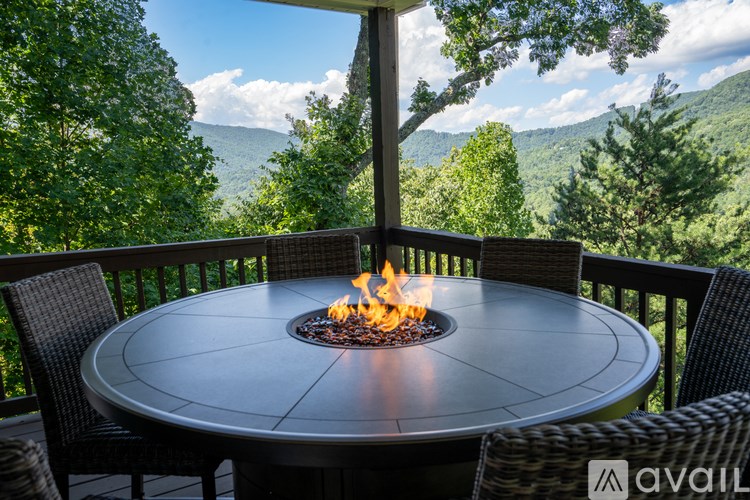A table with a fire pit in the middle is surrounded by chairs on a deck with a mountain view.