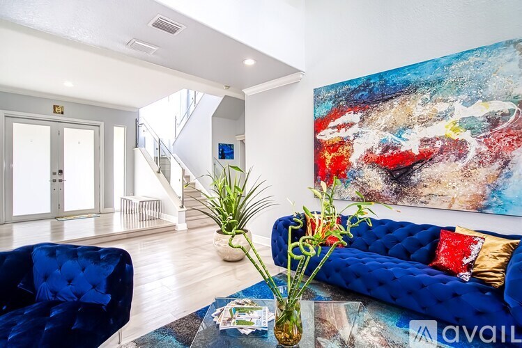 A blue couch with a red pillow is in the foreground of a bright room with a painting on the wall.