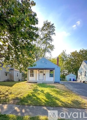 A small blue house with a white door is surrounded by greenery and other houses.