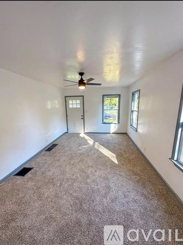 A room with a carpeted floor, a ceiling fan, and a door leading to another room.