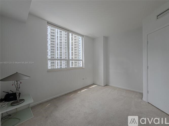 A room with a large window covered in blinds, a lamp on a table, and a carpeted floor.