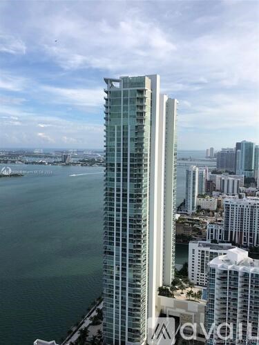A tall glass building stands in the foreground with a body of water and other buildings in the background.