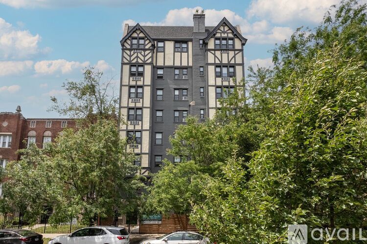A tall building with a mix of grey and white sections is surrounded by green trees and parked cars.