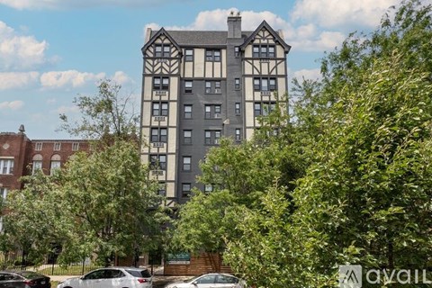 A tall building with a mix of grey and white sections is surrounded by green trees and parked cars.