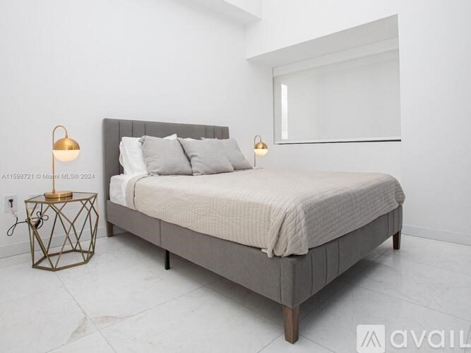 A bed with a grey headboard and a white bedspread is in a room with a white wall and a window.
