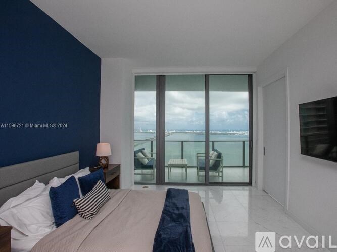 A bedroom with a bed, pillows, a lamp, and a large window overlooking a balcony.