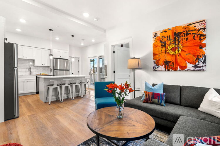 A modern kitchen with a dining area and a living room with a grey couch and a large orange painting.