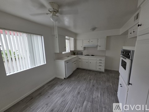A kitchen with white appliances and cabinets, a ceiling fan, and a window with blinds.