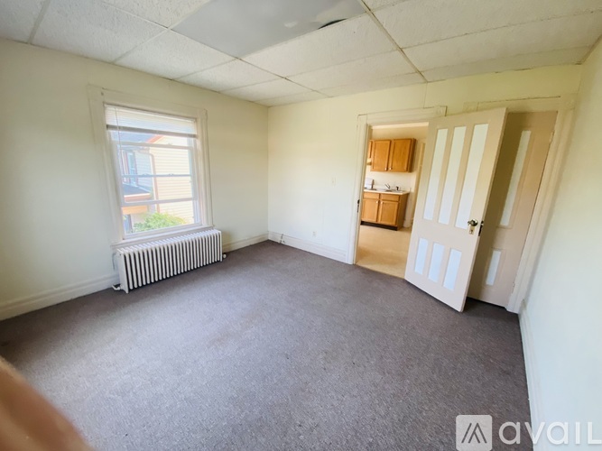 A room with a carpeted floor, a window with a view of a building outside, a white radiator, and a door leading to another room.