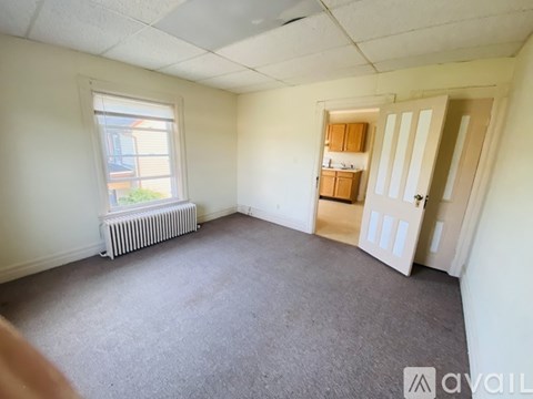 A room with a carpeted floor, a window with a view of a building outside, a white radiator, and a door leading to another room.