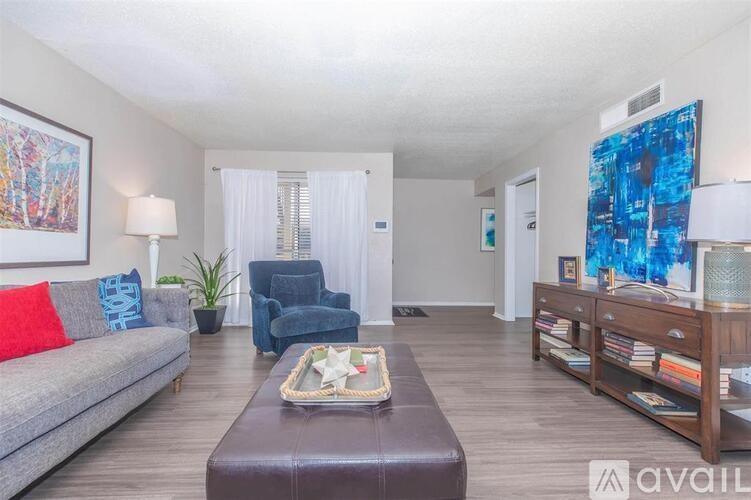 A living room with a grey couch, a brown leather ottoman, a blue armchair, a wooden bookshelf, and a painting on the wall.