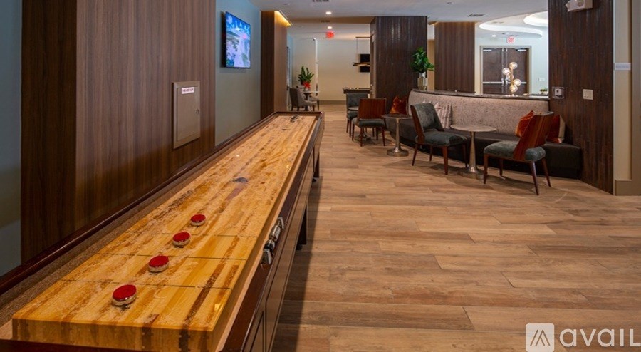 A long wooden table with red buttons in the middle is in the foreground of an indoor room with a TV and a bar in the background.