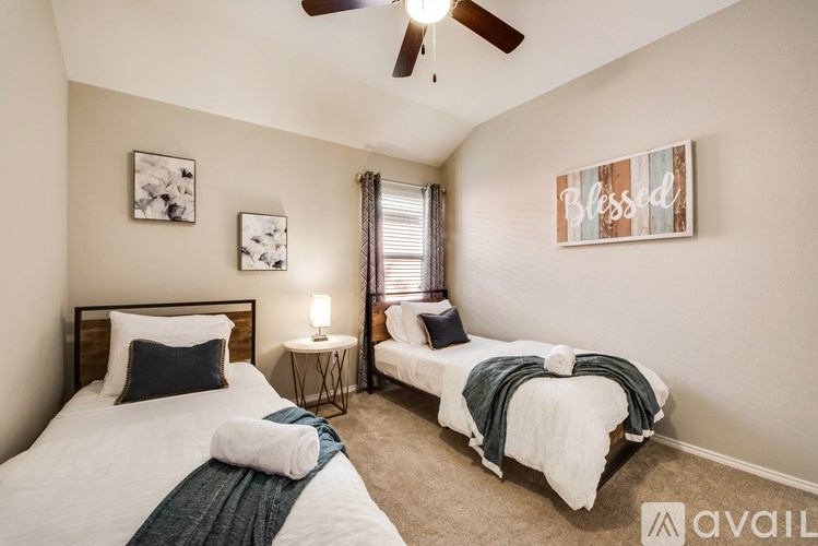 A bedroom with two beds, one with a white comforter and the other with a blue comforter, a ceiling fan, and a painting with the word "Blessed" on the wall.