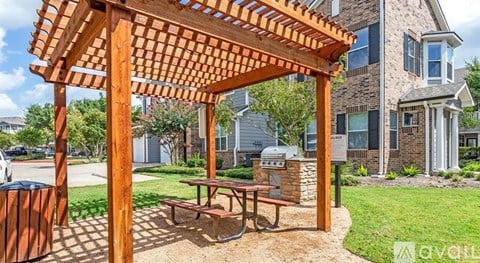 A wooden pergola with a picnic table is situated in a grassy area in front of a residential building.