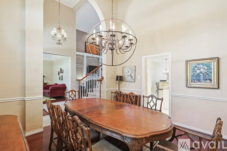 A large wooden dining table with chairs and a chandelier hangs in a room with a staircase in the background.