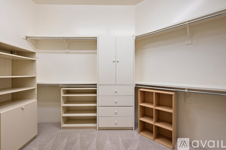 A room with white walls and a grey carpet has a white wardrobe with drawers and shelves, a white cupboard with shelves, and a white cabinet with shelves.