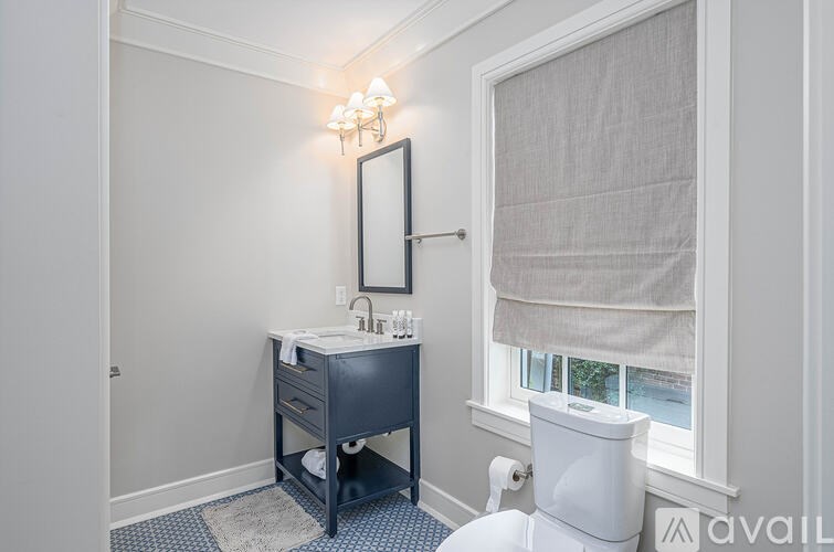 A bathroom with a white toilet, a sink with a mirror and a light above it, and a window with a curtain.