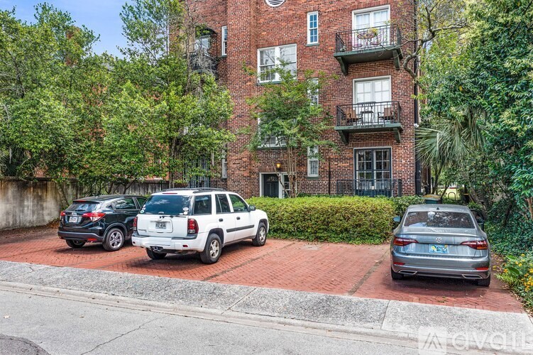 A red brick building with a balcony on the second floor is surrounded by trees and has cars parked in front.