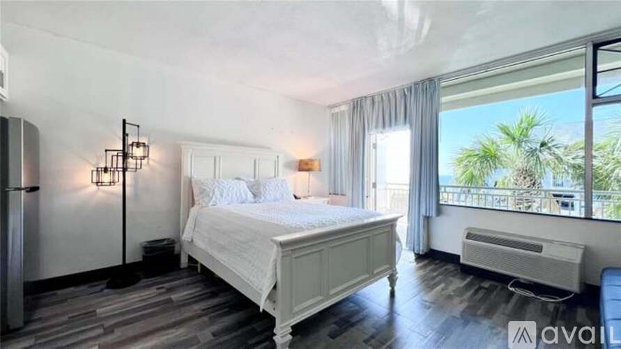 A bedroom with a large bed, a window with a view of palm trees, and a refrigerator.