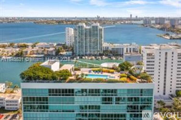 A modern glass building with a pool on the roof is surrounded by other buildings and a body of water.