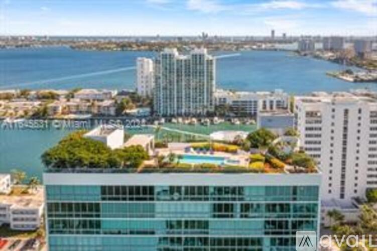 A modern glass building with a pool on the roof is surrounded by other buildings and a body of water.