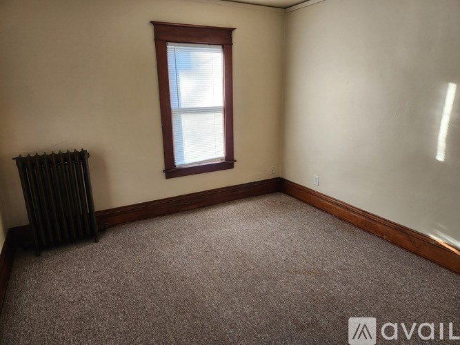 A room with a carpeted floor, a window with blinds, and a radiator.
