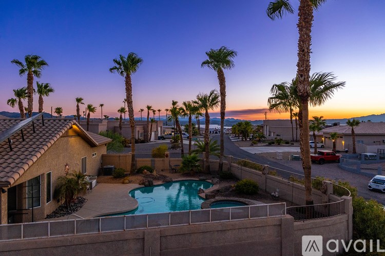 A sunset view of a pool surrounded by palm trees and a house with a car parked in front.