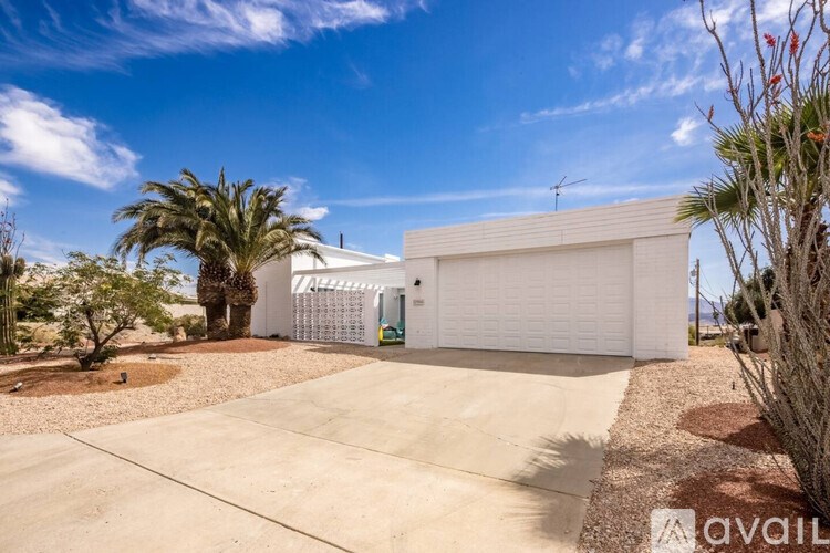 A white house with a garage door is surrounded by a gravel driveway and palm trees.