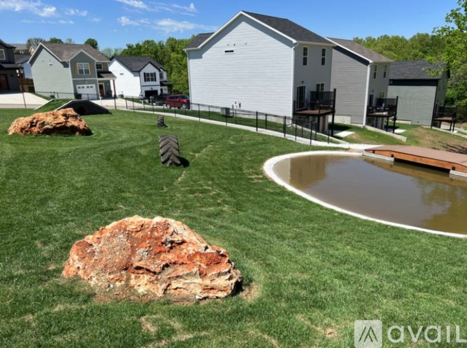 A large rock sits in the middle of a grassy area with a pond and houses in the background.