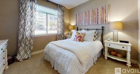 A bedroom with a white bed, a white nightstand, and a window with curtains.
