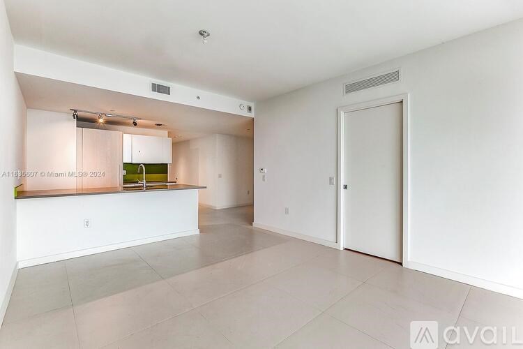 A spacious, empty room with white walls and flooring, and a kitchen area with a counter and cabinets in the background.