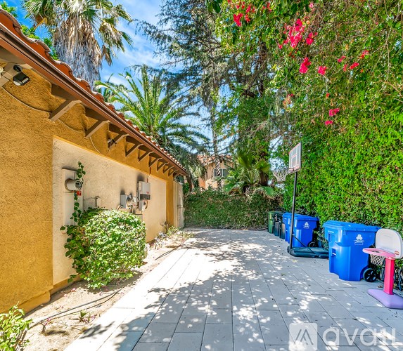 A sunny day in a narrow street with a yellow wall on the left and blue bins on the right.