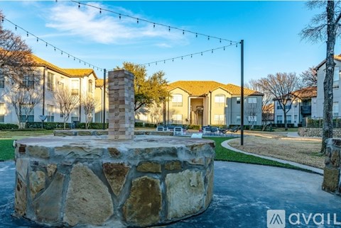 A stone fountain is in the middle of a grassy area with houses in the background.