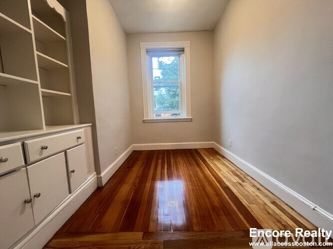 A room with wooden floors and white walls, with a window and a cabinet on the left side.
