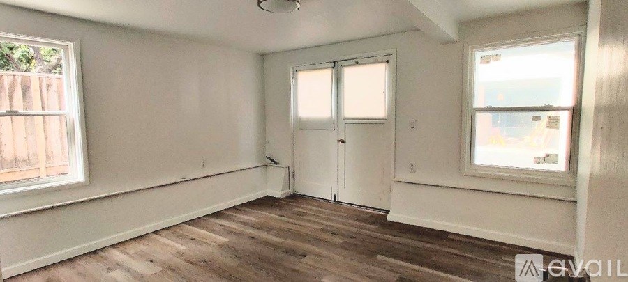 A room with wooden floors and white walls, featuring a window with a view of a fence and a door.