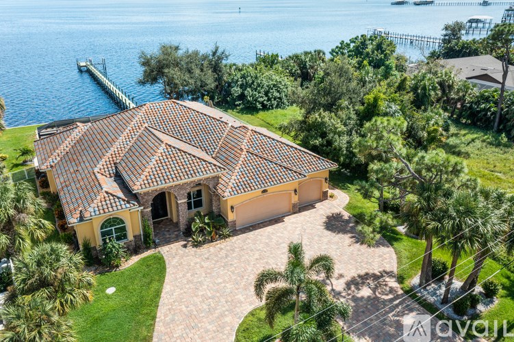 A house with a red tile roof is surrounded by greenery and a body of water.