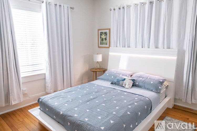 A bed with a blue comforter and white pillows is in a room with white walls and a window with white curtains.