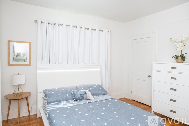 A bedroom with a bed, a nightstand, a lamp, a picture on the wall, and a vase of flowers on the dresser.