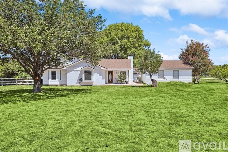 A white house with a brown roof is surrounded by a green lawn and trees.