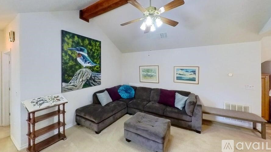 A living room with a grey couch, a painting of a bird, a side table, and a ceiling fan.