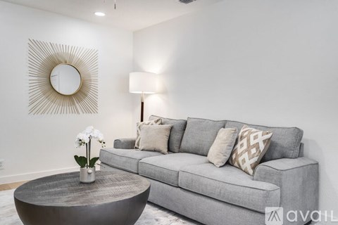 A living room with a grey couch, a round mirror, and a coffee table with a vase of flowers on it.