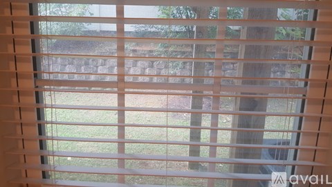 A window with white blinds partially open, allowing a view of a grassy area outside.