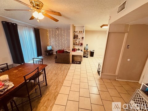 A living room with a dining table and chairs, a ceiling fan, and a kitchenette in the background.