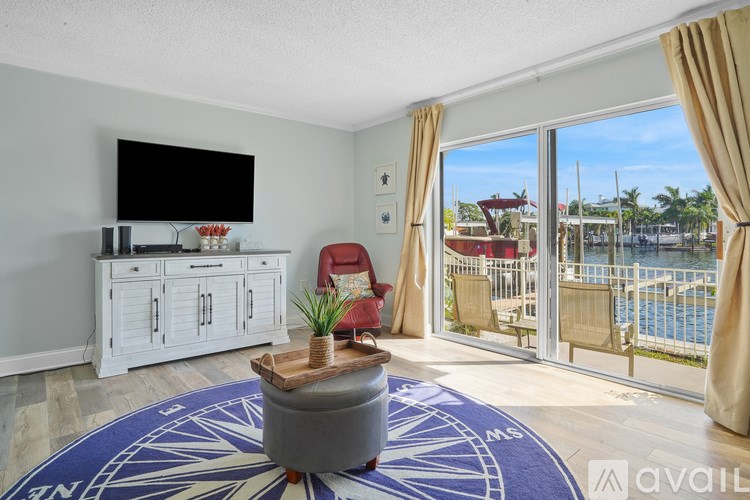A living room with a flat screen TV, a white cabinet, a grey ottoman, and a view of a pool through the sliding glass doors.