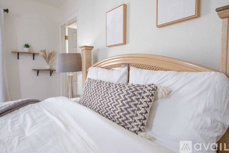 A bed with a white comforter and a brown and white pillow is in the foreground of a room with a lamp and two framed pictures on the wall.