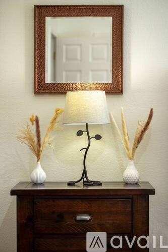 A brown framed mirror hangs on a wall above a wooden dresser with a lamp and two vases on top.