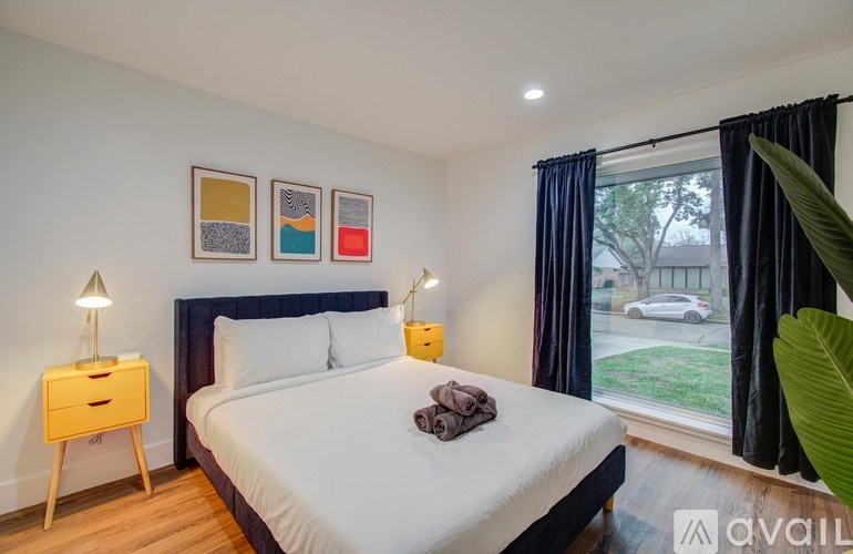 A bedroom with a large bed, a nightstand with a lamp, and a window with a view of a car and trees outside.