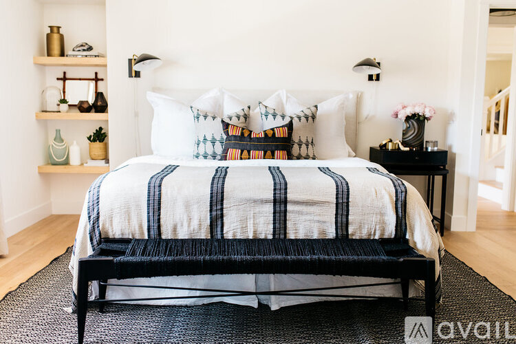 A bed with a white and blue striped comforter and pillows is in the foreground of a room with a black chair and a small table with a vase of flowers.