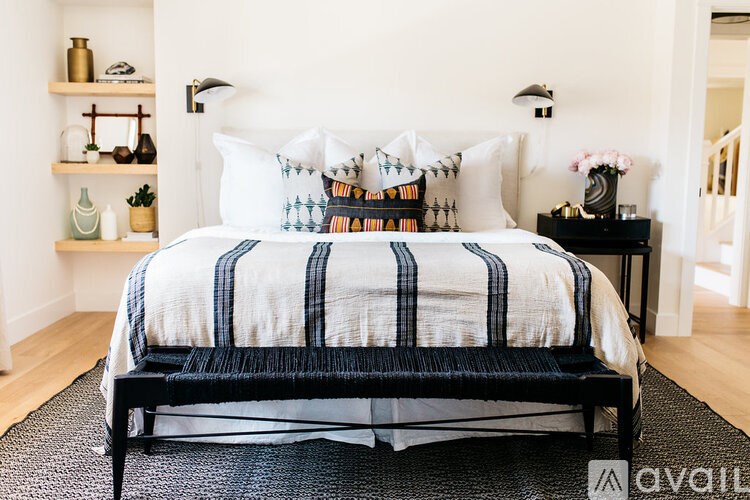 A bed with a white and blue striped comforter and pillows is in the foreground of a room with a black chair and a small table with a vase of flowers.